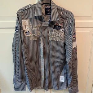 Randig skjorta från Camp David - Skit snygg blå och vit randig skjorta från Camp David med broderade detaljer och patchar på bröstet, ryggen och ärmarna. Skjortan har långa ärmar, klassisk krage och knäppning framtill. I strl M