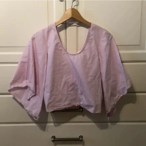 Ljusrosa croppad blus med vida ärmar från Zara - Säljer en ljusrosa croppad blus från Zara med vida trekvartsärmar och öppen rygg med rosett. Blusen är i bomull och har en lös passform, i strl xs