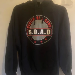Svart hoodie med System of a Down-tryck - Svart hoodie med stort tryck av System of a Down (S.O.A.D) på bröstet. Tröjan har huva med dragsko och magficka. Storlek Medium men passar som Small, hoodien är från tidigt 2000 tal