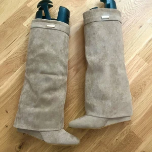 Beiga fold-over mockastövlar med kilklack - Helt oanvända!! Säljer ett par beiga fold over stövlar i mocka med kilklack och dragkedja på insidan. Köpta från lululia, slutsåld design för 799kr. Klack ca 7cm. Aldrig kommit till använding. Strl 38🫶