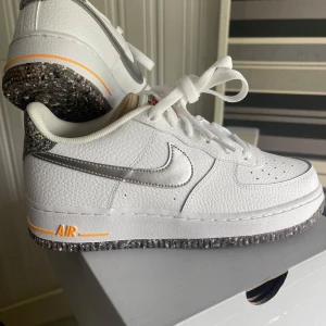 Nike Air Force 1 '07 GS White/Metallic Silver - Säljer ett par fräscha Nike Air Force 1 '07 GS i vitt med metallic silver swoosh och detaljer. Skorna har en unik gråspräcklig sula och orange AIR-tryck på sidan. Klassisk låg modell med snörning och perforerad tå för extra ventilation.