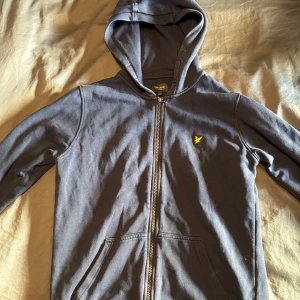 Mörkgrå hoodie med dragkedja från Lyle & Scott - Mörkgrå hoodie med dragkedja och huva från Lyle & Scott. Jackan har två fickor framtill och en gul broderad logga på bröstet. Perfekt för en avslappnad stil.