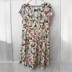 Blommig kortärmad klänning med v-ringning - Söt klänning med blommigt mönster i rosa, blått, gult och grönt på vit botten. Kortärmad modell med v-ringning och dekorativ öppning vid halsen. Klänningen har en lätt utställd kjol och är perfekt för varma dagar.