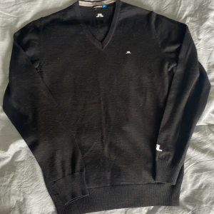 Svart v-ringad tröja från J.Lindeberg - Nypris 1400, Klassisk svart v-ringad tröja från J.Lindeberg med diskret logga på bröstet och nacken. Tröjan är ribbad vid ärmslut och nederkant och har en stilren design som passar till mycket. Tillverkad i merinoull, ett tunt och mjukt material. Två små hål som syns vid utsidan av etiketten, se bild 4.