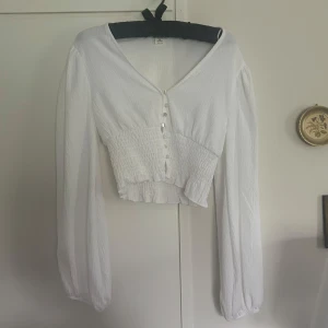 Vit blus med pärlemorknappar - Säljer en vit blus med långa ärmar och v-ringning. Blusen har smockad midja och pärlemorknappar framtill. Tunt, lätt material som ger en luftig känsla. Perfekt till vår och sommar!🥰 Defekter: saknar en knapp längst ner samt några små fläckar på ena armen.