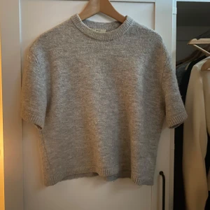 Grå stickad tröja från Gina Tricot - Mysig grå stickad tröja från Gina Tricot med korta ärmar och rund halsringning. Tröjan har en rak passform och ribbade kanter.