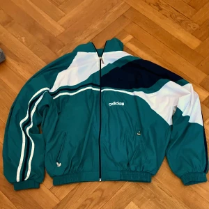 Grön och vit vindjacka från Adidas - Retro vindjacka från Adidas i grönt, vitt och mörkblått med dragkedja framtill och klassiska ränder. Jackan har hög krage, resår i midja och ärmslut samt två fickor med dragkedja. Perfekt för en sportig look. Sen är väldigt oversized och ska kunna passa L och M. Den är från 90-talet