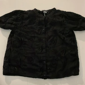Svart blus med broderade detaljer - Svart blus med korta ärmar och knappar framtill. Blusen har broderade hålmönster som ger en unik look och en lätt glansig yta. Perfekt för dig som vill ha något stilrent men ändå med det lilla extra.