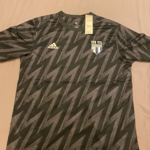 Svart och grå fotbollströja från Adidas - Säljer en svart fotbollströja från Adidas med grått blixtmönster och klubbmärke på bröstet där det står '120 YIL'. Tröjan har korta ärmar och rund hals. Perfekt för dig som gillar unik design och sportig stil.