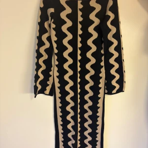 Svart och beige långklänning från NA-KD - Säljer en långklänning från NA-KD i svart med beige vågigt mönster. Klänningen har lång ärm och är stickad i mjukt material. Perfekt för dig som gillar unika mönster och vill sticka ut.