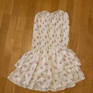 Blommig axelbandslös klänning med volang - Söt axelbandslös klänning i ljus färg med små rosa blommor och volangkjol. Smockad överdel ger en bekväm passform och kjolen har flera lager för extra volym. Perfekt för sommarens varma dagar.