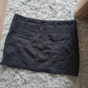 Svart kortkjol med fickor - Säljer en svart kortkjol med två fickor framtill och knappstängning i midjan. Kjolen har en enkel och stilren design som passar till många olika outfits. Perfekt för dig som gillar en klassisk look.