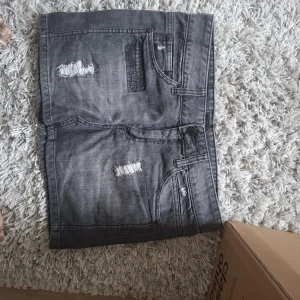 Grå jeanskjol med slitningar - Säljer en grå jeanskjol med coola slitningar framtill och baktill. Kjolen har klassiska fickor både fram och bak samt knapp och dragkedja i midjan. Perfekt för dig som gillar en edgy stil.