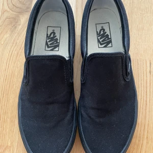 Svarta Vans Classic Slip-On - Säljer ett par svarta Vans Classic Slip-On med helsvart canvas och sula i storlek 40. Skorna har en enkel och stilren design utan snörning. Kommer i originalkartong. Använda en gång och är i mycket fint skick.