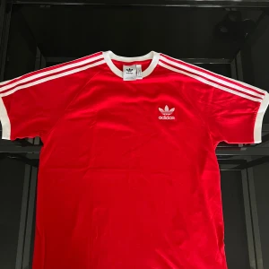 Röd t-shirt från Adidas Originals - Säljer en klassisk röd t-shirt från Adidas Originals med vita ränder på axlarna och ärmarna samt vit logga på bröstet. T-shirten har rund halsringning och korta ärmar. Perfekt för en sportig look!