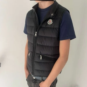 Svart dunväst från Moncler - Säljer en svart dunväst från Moncler med klassisk logotyp på bröstet. Västen har hög krage, två fickor med dragkedja och quiltad design. Perfekt för lager-på-lager och stilren look. Stolek 152 12 år. Skriv om ni har frågor🔥🔥🔥🔥🔥