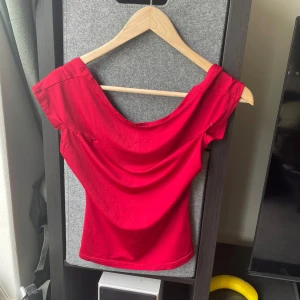Röd croppad topp med knutdetalj - Säljer en röd croppad topp med offshoulder-design och snygg knutdetalj framtill. Toppen har en figurnära passform och är tillverkad i ett stretchigt syntetmaterial. Hål vid ärmarna