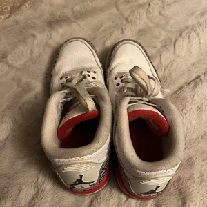 Nike Air Jordans vita med röd och grå detalj - Säljer ett par klassiska Nike Air Jordans i vitt skinn med grå och svarta mönstrade detaljer samt röda inslag. Skorna har snörning och en rund tå. Perfekta för dig som gillar streetwear och sneakers med ikonisk design.