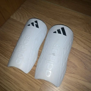 Vita benskydd från Adidas - Säljer ett par vita benskydd från Adidas med svart logga upptill. De har en stilren design med diskret mönster längs sidorna och är gjorda i ett tåligt plastmaterial. Perfekta för fotbollsträning eller match.
