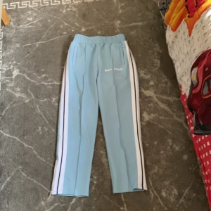 Ljusblåa träningsbyxor från Palm Angels - Snygga ljusblåa träningsbyxor från Palm Angels med vita och svarta ränder längs sidorna. Byxorna har resår i midjan och dragkedja vid benslut. Perfekta för en sportig och trendig look.