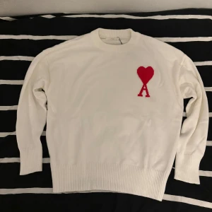 Vit stickad tröja från Ami Paris med rött hjärta - Säljer en vit stickad tröja från Ami Paris med ett rött hjärta och bokstaven A broderat på bröstet. Tröjan har rund halsringning och ribbade muddar vid ärmslut och nederkant. Perfekt för dig som gillar stilrena plagg med en twist. Kontakta för mer information.