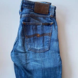 Blå jeans från Nudie Jeans - Säljer ett par klassiska blå jeans från Nudie Jeans med fem fickor och orangea sömmar. Jeansen har normal passform och raka ben, tillverkade i bomull. Perfekta för dig som gillar en tidlös stil.
