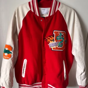Röd och vit collegejacka med broderier från Bershka - Säljer en röd och vit bomberjacka från Bershka med broderade detaljer som en grön drake, eld och texten 'COOLEST' på ryggen. Jackan har knappar framtill, ribbade muddar och två fickor. Perfekt statement-plagg för dig som vill sticka ut. Där är en liten missfärgning på ena ärmen.