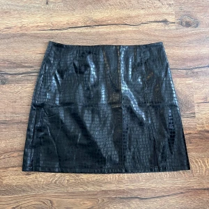 Svart kjol i skinnimitation med ormmönster - Snygg svart kjol i glansig skinnimitation med ormmönstrad struktur. Rak modell med kort längd.