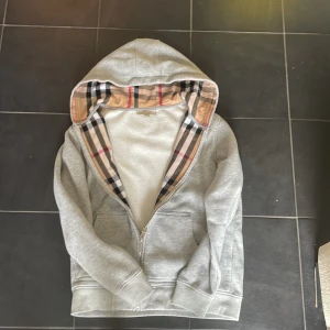 Grå hoodie från Burberry med klassiskt rutigt foder - Säljer en grå hoodie från Burberry med dragkedja och två fickor framtill. Insidan av huvan har det ikoniska Burberry-rutiga fodret i beige, svart och rött. Perfekt för en stilren och avslappnad look.