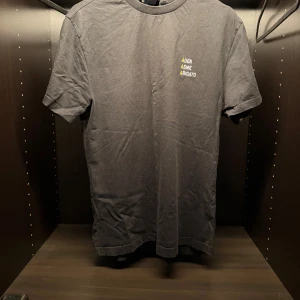 Svart t-shirt från Axel Arigato - Svart t-shirt från Axel Arigato med tryck i vitt och gult på bröstet där det står 'ADEN ASME ARIGATO'. Klassisk rund hals och korta ärmar. Perfekt för en clean och stilren look.
