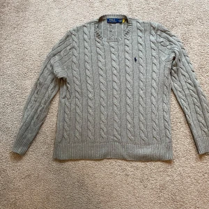 Grå kabelstickad tröja från Polo Ralph Lauren - Säljer en klassisk grå kabelstickad tröja från Polo Ralph Lauren med rund halsringning och broderad logga på bröstet. Tröjan har långa ärmar och ribbade muddar vid ärmslut och nederkant. Perfekt för lager-på-lager-stil.