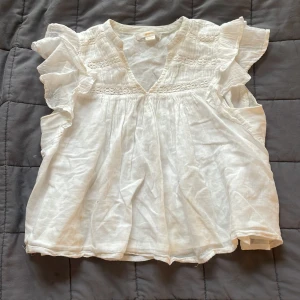 Vit blus med volang från Gina Tricot - Säljer en söt vit blus från Gina Tricot med volangdetaljer på axlarna och v-ringning. Blusen har korta ärmar och är tillverkad i ett lätt och luftigt material, perfekt för varmare dagar.