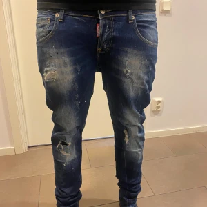 Blå jeans från Dsquared2 med slitningar - Säljer ett par blå jeans från Dsquared2 med tydliga slitningar och färgstänk på framsidan. Jeansen har normal passform och klassisk femficksdesign. Stor Dsquared2-logga på bakfickan och läderpatch i midjan.
