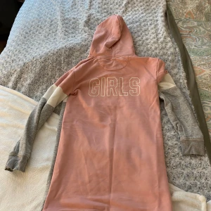 Rosa och grå hoodie från GANT - Säljer en snygg hoodie från GANT i rosa och grått med vit detalj och texten 'GANT' framtill och 'GIRLS' på ryggen. Tröjan har huva och långa ärmar, perfekt för en avslappnad stil.
