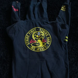 Svart Cobra Kai hoodie från Ellesse storlek M - Svart hoodie från Ellesse med Cobra Kai-tryck på ryggen och texten 'Strike First, Strike Hard, No Mercy' i gult. Röd Ellesse-logga på ena ärmen och gula detaljer på den andra. Klassisk huva och långa ärmar, perfekt för dig som gillar streetstyle och serier.