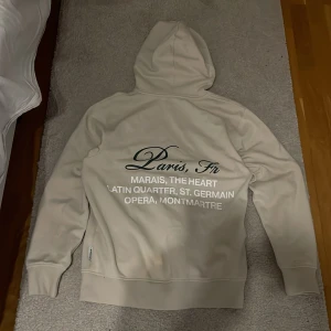 Beige hoodie med Paris-tryck - Snygg beige hoodie med tryck på både bröstet och ryggen där det står 'Paris, Fr' och namn på olika stadsdelar i Paris. Hoodien har huva, långa ärmar och ribbade muddar. Perfekt för dig som gillar stilrena plagg med en touch av storstadsvibe. Det finns några små fläckar men inget som man tänker på.