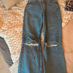 Blå ripped jeans från Pull&Bear - Säljer ett par blå jeans från Pull&Bear i klassisk denim med snygga slitningar över knäna. Modellen är raka och har hög midja, perfekt för en avslappnad och trendig look. Jeansen har fem fickor och dragkedja med knapp. Bra skick. 