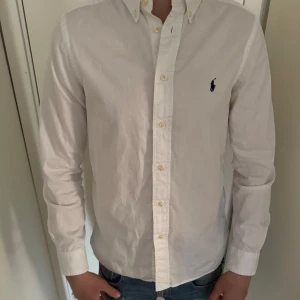 Vit skjorta från Polo Ralph Lauren - Klassisk vit skjorta från Polo Ralph Lauren i slim fit-modell. Skjortan har lång ärm, knappar framtill och den ikoniska broderade loggan på bröstet. Tillverkad i mjuk bomull och passar perfekt till en clean och stilren look.