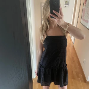 Svart ärmlös kortklänning med volang - Supersöt svart kortklänning med tunna axelband och volang nertill. Klänningen har en rak passform och är sydd i ett broderat tyg med diskret mönster. Perfekt för dig som gillar en enkel men ändå detaljerad look.