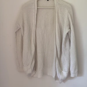 Vit stickad kofta från Bik Bok - Mysig vit stickad kofta från Bik Bok i 100% bomull. Koftan har långärmad design, öppen framtill och två praktiska fickor. Perfekt för lager-på-lager och chill vibes. Ribbstickade muddar och avslappnad passform gör den extra comfy.