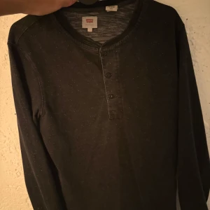 Svart långärmad tröja från Levi's - Svart långärmad tröja från Levi's med diskreta vita prickar och knappar vid halsen. Tröjan har en avslappnad passform och är perfekt för dig som gillar en enkel men stilren look. Tillverkad i mjuk bomull för extra komfort.