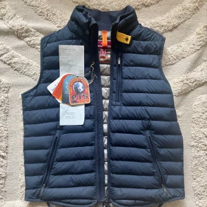 Mörkblå dunväst från Parajumpers - Snygg mörkblå dunväst från Parajumpers med quiltad design och hög krage. Västen har dragkedja framtill, två fickor med dragkedja och en gul detalj vid halsen. Klassisk PJS-logga på ryggen. Perfekt för lager-på-lager under kyliga dagar.