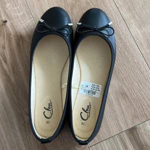Svarta ballerinaskor med rosett från Clou - Klassiska svarta ballerinaskor från Clou i storlek 38. Skorna har rund tå, platt sula och en söt rosettdetalj framtill med silvriga ändar. Ytan är slät och materialet känns syntetiskt. Perfekta för en stilren och enkel look. Helt nya och aldrig använda. Pris kan diskuteras 😊