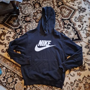 Svart Nike hoodie med stor logga - Svart hoodie från Nike med stor vit logga framtill. Klassisk känguruficka, huva med snörning och långa ärmar. Perfekt för dig som gillar sportig och avslappnad stil. Mjuk bomullsblandning som är skön att ha på sig hela dagen.