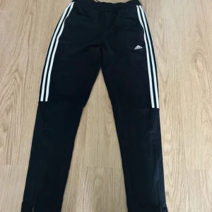 Svarta Adidas mjukisbyxor med ränder - Svarta mjukisbyxor från Adidas med klassiska vita ränder längs sidorna och liten Adidas-logga på låret. Byxorna har elastisk midja och är tillverkade i ett mjukt och bekvämt material, perfekta för träning eller chill. Snygg och sportig look.