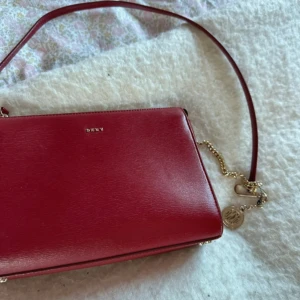 Röd axelväska från DKNY  - ❤️Snygg röd axelväska från DKNY i slätt skinn med guld detaljer och kedja. Väskan har en stilren med DKNY-logo framtill. Perfekt storlek för det viktigaste och en klassisk look med exklusiv känsla.❤️
