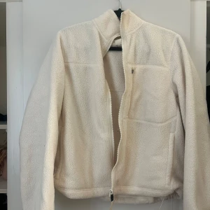 Beige teddyjacka med dragkedja - Mysig beige teddyjacka med hög krage och hel dragkedja framtill. Jackan har en bröstficka med dragkedja, två sidofickor och justerbar dragsko i nederkant. Perfekt för kalla dagar när du vill vara både varm och snygg.