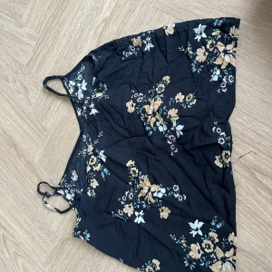 Blommig svart linnetopp från Divided - Svart linnetopp från Divided med tunna axelband och blommigt mönster i beige, vitt och grönt. Toppen är luftig och perfekt för varma dagar, med en avslappnad passform och mjukt material. Snygg att matcha med jeans eller kjol för en chill look.