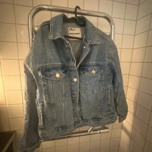 Blå jeansjacka från Sister Point - Klassisk blå jeansjacka från Sister Point med silverfärgade knappar och två bröstfickor med lock. Jackan har en rak passform, krage och långa ärmar. Perfekt att slänga över en hoodie eller t-shirt för en chill look.