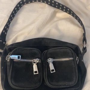 Svart axelväska  - Cool svart axelväska med bred axelrem dekorerad med silvriga nitar. Väskan har två stora framfickor med tydliga silvriga dragkedjor och är gjord i ett mjukt tygmaterial. Perfekt för att få plats med allt du behöver till vardagen.